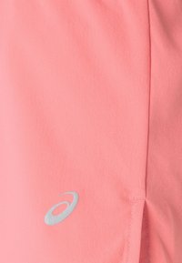 Vaaleanpunaiset shortsit, tehty pehmeästä kankaasta, joissa on hopeinen logo ja sivuhalkiot liikkuvuutta varten. Sileä pinta ja siistit ompelut.