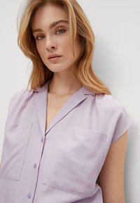 Light lavender short-sleeve blouse with a collar, featuring two chest pockets and button detail, made from a smooth fabric.