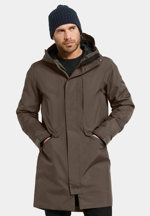 Brown waterproof coat with a hood, two front pockets, and adjustable cuffs. Paired with a dark ribbed knit beanie.