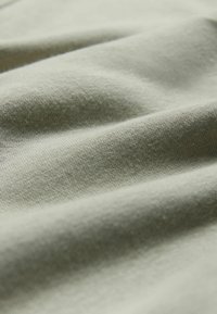 Vaaleanvihreä puuvillakangas, jossa on sileä pinta, hienovaraiset taitokset ja pehmeä, hengittävä ilme. Lähikuva yksityiskohdasta.