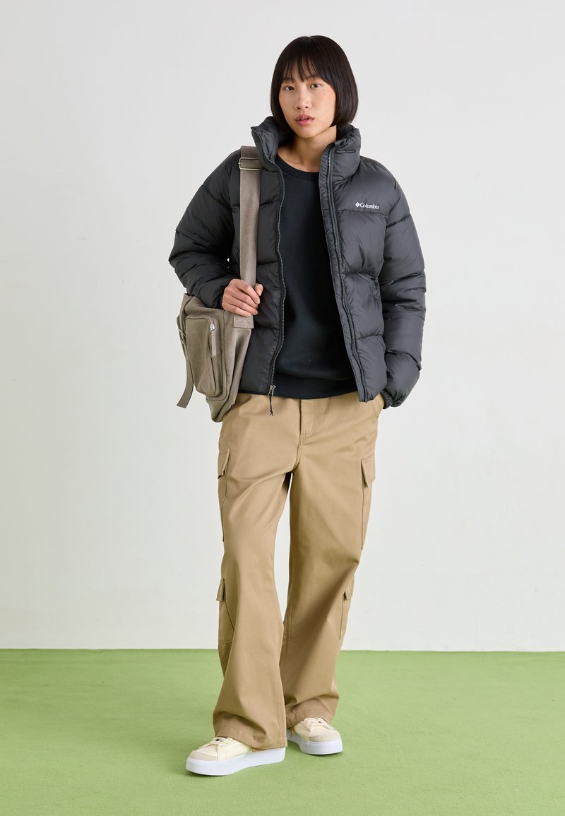 Zwarte pufferjack, zwarte sweatshirt, beige cargobroek en witte sneakers. Het model houdt een tan schoudertas vast met een ritszak.