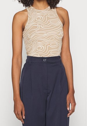 Femme portant un haut beige sans manches avec des lignes blanches abstraites et un pantalon taille haute bleu marine plissé.