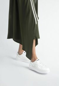 Sneakers bianchi in pelle liscia, dotati di occhielli e di un dettaglio con logo. Abbinati a un vestito verde oliva con strisce laterali bianche.