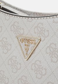 Beige-texturiertes Stoffhandtasche mit einem goldenen dreieckigen Logo, auf dem "GUESS" und ein Fragezeichen zu sehen sind. Dezente gemusterte Oberflächendetails sind sichtbar.