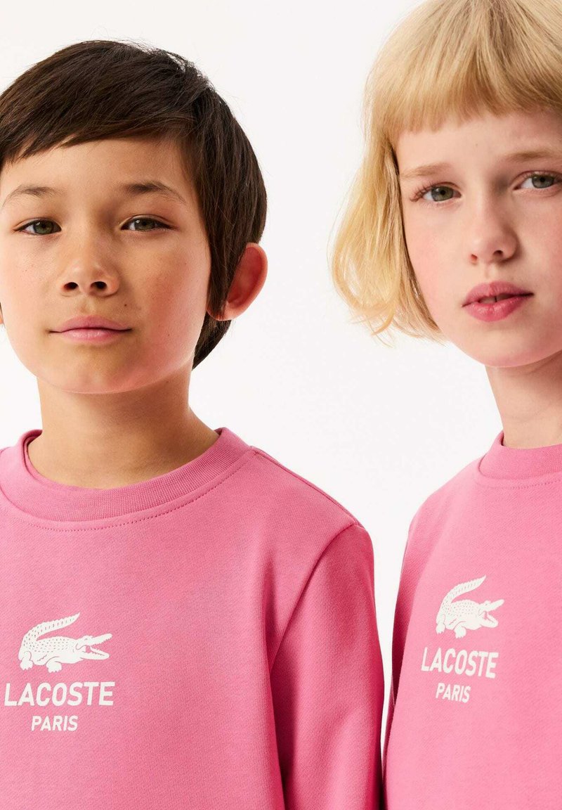 Rosa Baumwoll-Sweatshirts mit weißem Lacoste-Logo. Rundhalsausschnitt, gerippte Bündchen, glatte Textur, minimalistischem Stil. Nahaufnahme von zwei Modellen.