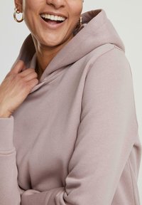 Hoodie mauve clair avec une texture douce, doté d'une grande capuche et d'une coupe décontractée. Le modèle a une main près de l'encolure, mettant en valeur le tissu.