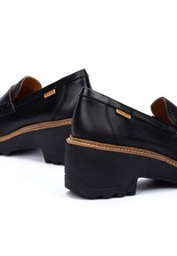 Zapatos negros de cuero tipo mocasín con una suela gruesa y texturizada y acentos marrones contrastantes alrededor de la entresuela. Etiqueta de logo visible en el lado.