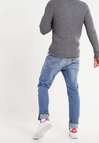 Osoba v šedém pleteném svetru, s modrými džínami s manžetami a bílými teniskami s červenými akcenty, stojící zády.