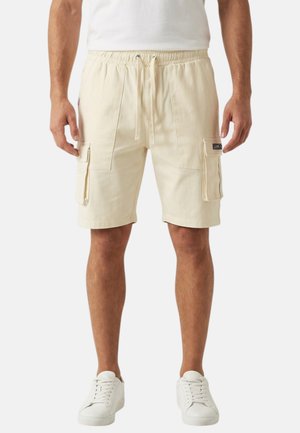 Homme portant un short cargo beige avec poches avant et latérales, des baskets blanches et un t-shirt blanc, debout devant un fond uni.