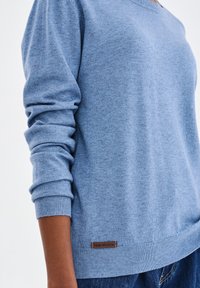 Jasnoniebieski sweter z długimi rękawami, prążkowanymi mankietami i dekoltem w kształcie litery V. Posiada brązową metkę z napisem "blue shadow" na dolnym brzegu. Miękka tekstura.