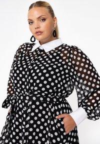 Schwarzes Kleid mit weißen Polka-Dots, durchsichtigen langen Ärmeln, einem weißen Kragen und Gürteldetails. Verfügt über Taschen und ein lockeres, fließendes Design.