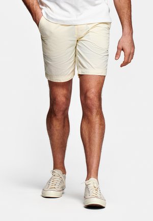 Man in lichtgele korte broek, witte sneakers en wit shirt met handen in de zakken, staand tegen een effen achtergrond.