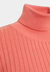 Pull à col roulé côtelé de couleur corail, présentant une texture douce et une coupe ajustée qui épouse délicatement le contour du cou.