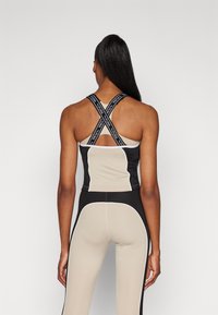 Top di abbigliamento attivo nero e beige con spalline incrociate, dotato di uno schienale con aperture e pannelli eleganti. I materiali sembrano lisci e elasticizzati.
