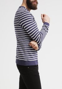 Camisa de manga larga a rayas en azul marino y blanco, con cuello redondo, diseño ajustado y puños acanalados. Lleva puesta sobre pantalones negros.