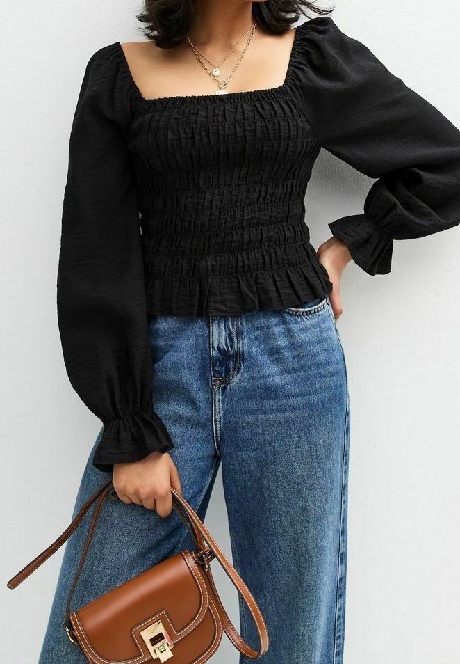 Femme portant un haut noir froncé à manches longues et un jean bleu, tenant un sac à main en cuir marron avec une fermeture dorée.