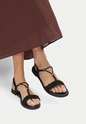 Sandale negre din suede cu design T-strap, având accente metalice aurii. Talpa plată cu vârfuri deschise și curele ajustabile la glezne.