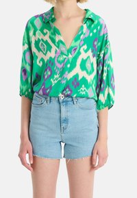 Zielona i fioletowa bluzka w wzory z dekoltem w kształcie litery V i luźnym krojem, połączona z jasnoniebieskimi szortami dżinsowymi z postrzępionymi brzegami.