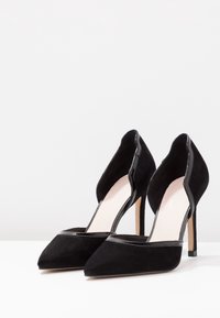 Escarpins en daim noir avec bouts pointus, bords festonnés et accents noirs brillants sur la ligne supérieure. Des talons aiguilles fins élèvent le design.