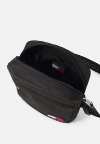 Borsa in nylon nera con zip superiore, dotata di una piccola tasca interna e un'etichetta con logo decorata con accenti rossi, bianchi e blu. Include una tracolla regolabile.