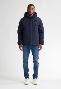 Laivastonsininen toppatakki hupulla, jossa edessä vetoketju ja taskut. Yhdistetty vaaleansinisiin farkkuihin ja valkoisiin lenkkareihin.