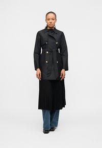 Άτομο με πλεγμένα μαλλιά που φοράει μαύρο παλτό τύπου trench με ζώνη πάνω από μαύρη πλισέ φούστα και μπλε τζιν, στέκεται μπροστά σε απλό φόντο.