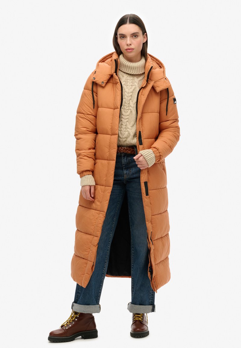 Lange oranje pufferjas met een capuchon, voorzien van gestikte secties, zwarte ritsen en verstelbare manchetten. Draagt over een crèmekleurige kabelgebreide trui en jeans.