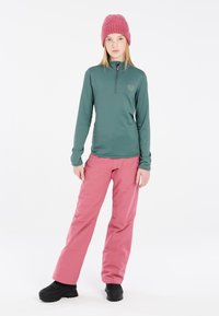 Langarm, groene thermoshirt met een half-rits kraag; roze, waterafstotende sneeuwbroek; zwarte, gestructureerde winterlaarzen; gebreide roze muts.