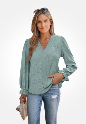 Blouse teal clair à manches longues avec décolleté en V, motif en œillets texturé et manches froncées. Assortie avec un denim usé et tenant une pochette.