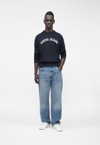 Navy džemperis ar uzrakstu "GUESS JEANS" baltā krāsā, kombinēts ar gaiši zilām plaša piegriezuma džinsa biksēm un baltām kedām. Vienkāršs fons.