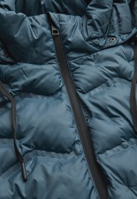 Mørkeblå pufferjakke med lynlås, tekstureret quiltning, elastiske snore og sorte detaljer langs lynlåsen og hætten.