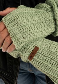 Muntgroene gebreide vingerloze handschoenen met een textuurpatroon en een leren label in bruin. Het materiaal ziet er dik en comfortabel uit.