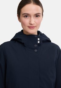Navy blue waterproof jacket with a high collar, button closures, and a drawstring hood, made from smooth, lightweight fabric.