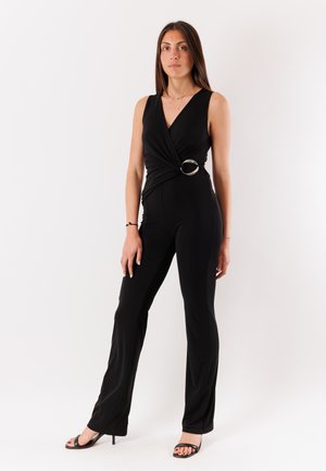 Donna in piedi, indossa una tuta nera senza maniche con dettaglio incrociato e anello d’argento in vita, abbinata a tacchi neri aperti in punta su sfondo bianco.