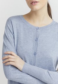 Cardigan bleu clair à manches longues en tissu doux, avec un col rond et cinq boutons sur le devant. Poignets côtelés.