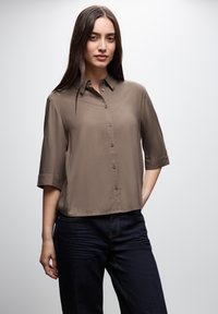 Camicia a maniche corte con bottoni in tessuto taupe. Presenta una vestibilità comoda, colletto e una trama liscia. Abbinata a jeans scuri.