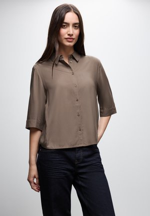 Chemise à manches courtes et à boutons en tissu taupe. Présente une coupe décontractée, un col et une texture lisse. Associée à un jean noir.