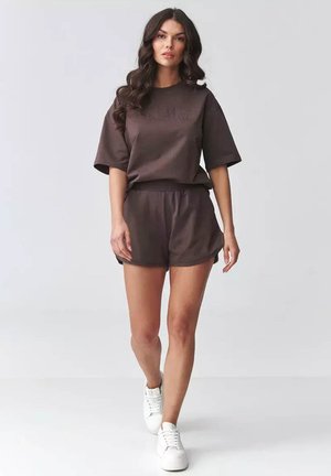 Kobieta idąca, ubrana w ciemnobrązowy, oversize top z krótkim rękawem i pasujące szorty oraz białe trampki na jednolitym tle.