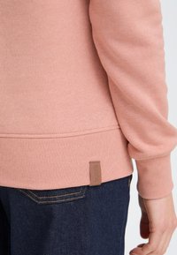Sudadera de color rosa claro con puños y dobladillo acanalados; presenta una tela texturizada y una pequeña etiqueta de cuero con marca en la parte inferior.