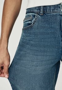 Strakke blauwe spijkerbroek met een zachte textuur, een voork pocket, zichtbare stikseldetails en een lichte stretch wanneer de stof wordt uitgerekt.