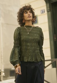 Eine Frau mit lockigem Haar trägt eine dunkelgrüne geraffte Bluse, übereinandergelegte Halsketten, auffällige Ohrringe und dunkle hochtaillierte Hosen im Freien.