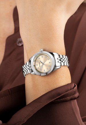 Montre en acier inoxydable argenté avec un cadran doré, des marqueurs d'heure en diamant et une lunette cannelée sur un bracelet à carreaux. Fond en tissu brun.