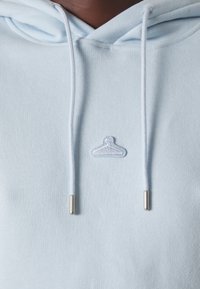 Sweat à capuche bleu clair avec cordons blancs et un petit symbole de cintre blanc brodé centré sur la poitrine.