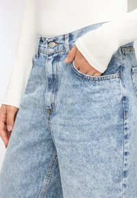 Jeans di denim azzurro chiaro a vita alta con effetto used, tasca frontale e chiusura con bottone metallico. Tessuto liscio e vestibilità ampia.