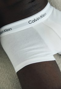 Weiße Baumwoll-Boxershorts mit einem schwarz-weißen Calvin Klein-Bund; glatte Textur. Auf einer hellen, strukturierten Oberfläche platziert.