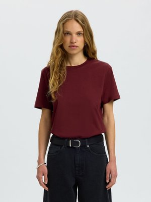 Ung kvinde med langt, bølget blond hår iført en ensfarvet vinrød t-shirt, sort bælte og sorte jeans mod en ensfarvet lys baggrund.