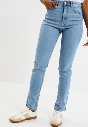 Frau trägt hellblaue Skinny-Jeans und weiße Sneakers mit beigen Akzenten, steht vor einem einfarbigen weißen Hintergrund.