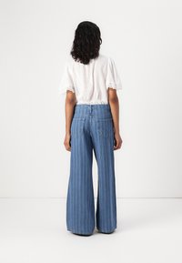 Blusa bianca a maniche corte con dettagli a occhio nei polsini, abbinata a jeans in denim leggeri a gamba larga di colore blu chiaro con righe verticali e tasche posteriori.
