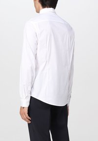 Camisa blanca de manga larga con una textura suave, cuello clásico y puños abotonados, con un dobladillo redondeado. Llevada con pantalones oscuros.