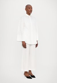 Camicia bianca oversize con bottoni e maniche larghe, abbinata a pantaloni bianchi a gamba larga e scarpe con tacco nere. Tessuto morbido, design minimalista.